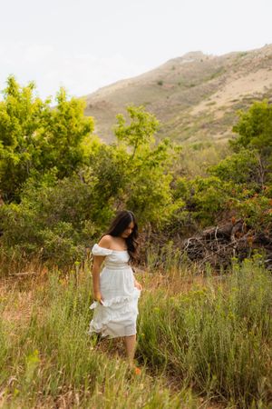 A figure in a flowy white dress walks through tall grass in a mountainous landscape with golden sunlight.
