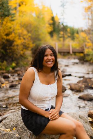 Smiling person sits on a rock by a stream surrounded by vibrant yellow fall foliage in a forest setting.