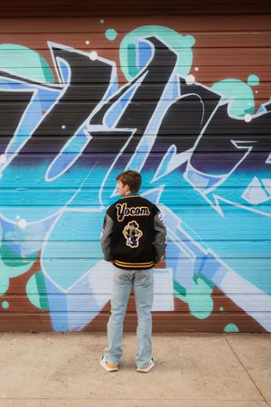 A person in a varsity jacket stands in front of a vibrant blue and turquoise graffiti mural on a garage door.