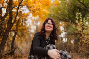 Person in glasses smiling and sitting outdoors among autumn trees with bright golden foliage in background.