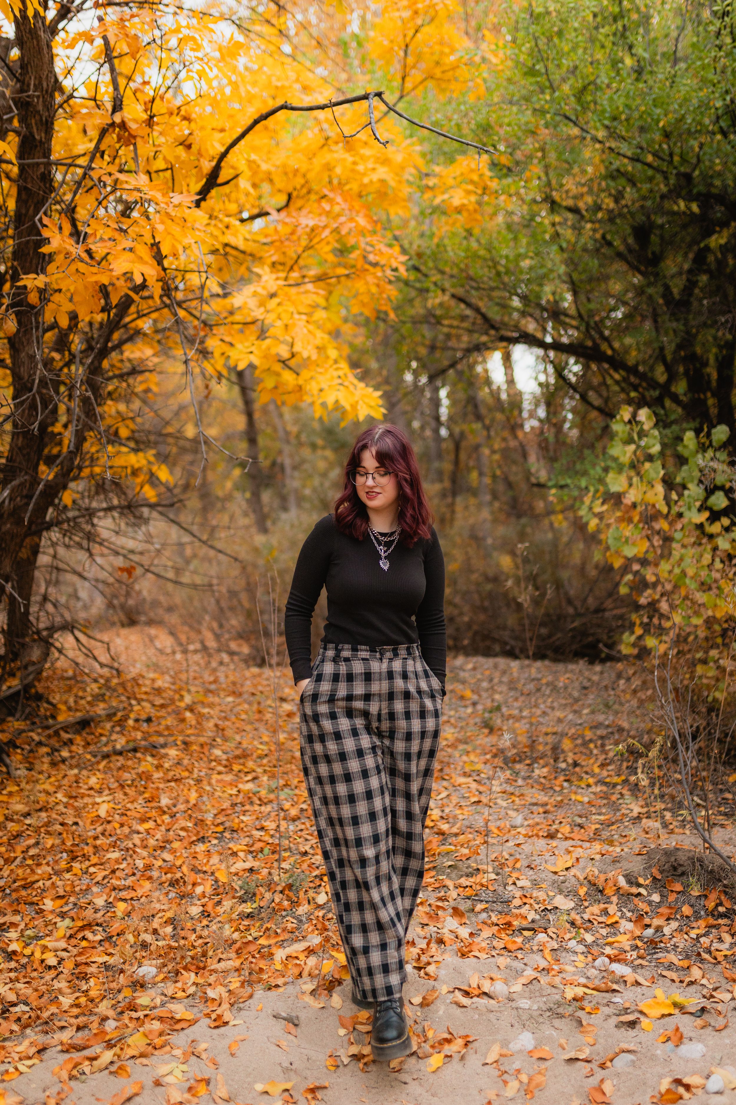 A figure in a plaid dress walks along a pathway covered in orange autumn leaves beneath golden trees in a park.