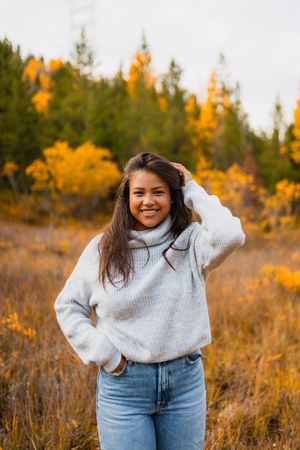 A person in a cozy white sweater and jeans poses outdoors against a backdrop of autumn trees with golden foliage.