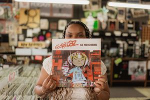 A person holds up a Gorillaz record album while browsing in a music store with shelves of vinyl records in the background.