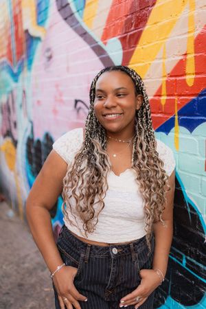 Person with long ombre braids wearing white top and striped pants poses against colorful graffiti mural wall.