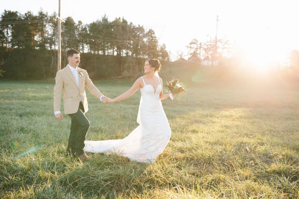 Couple holds hands while walking through sunlit meadow in wedding attire.