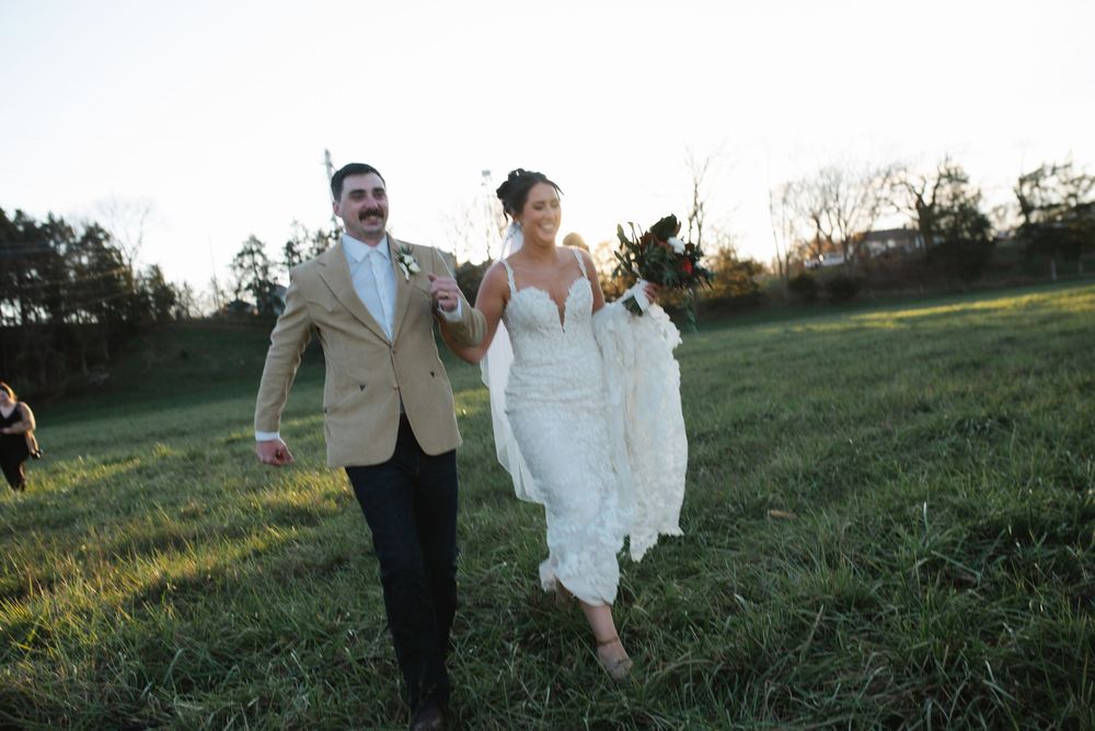 Newlyweds walking hand in hand through a sunlit field in formal attire.