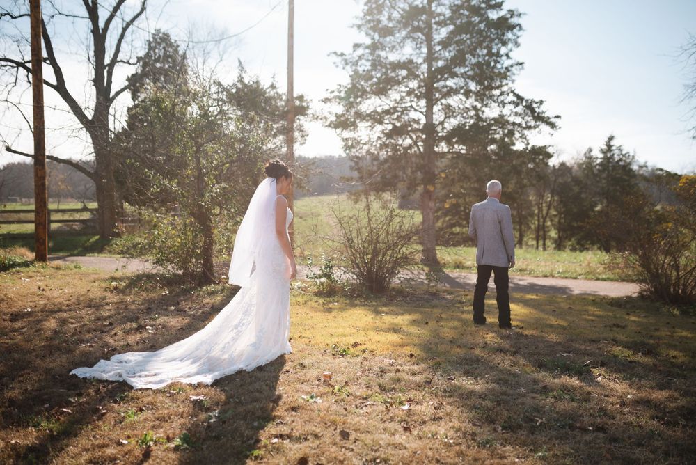 Bride in flowing white gown and groom stand apart in sun-dappled woodland setting.