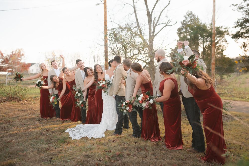 Wedding party poses casually outdoors with burgundy dresses and white flowers.