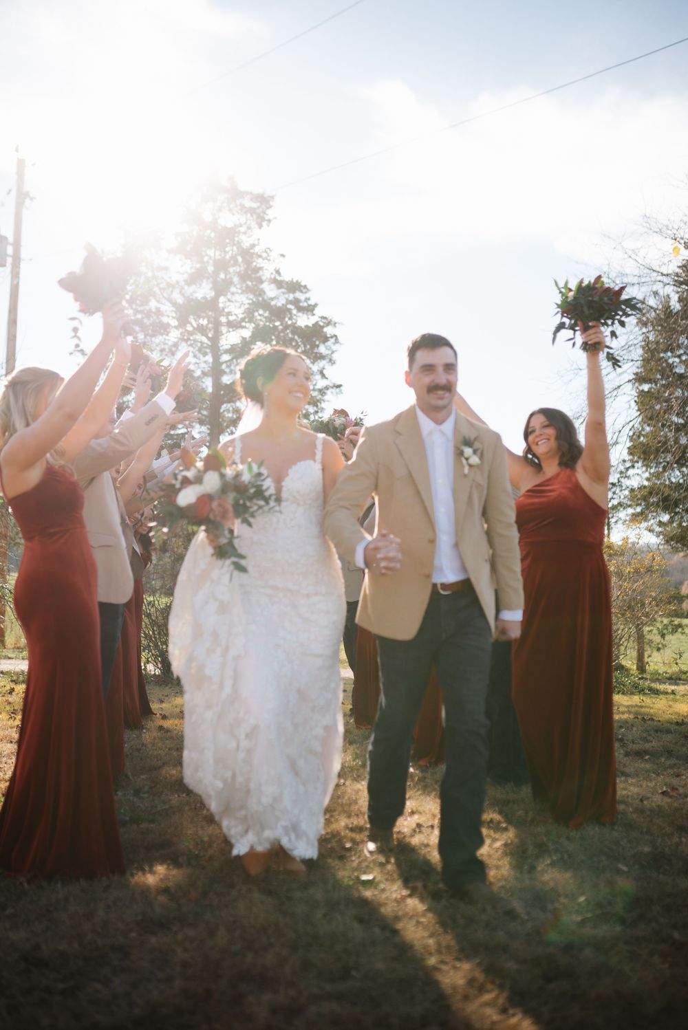 Wedding party celebrates with raised arms in burgundy dresses during golden hour.