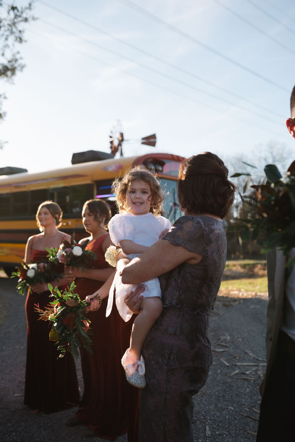 Wedding party members in burgundy dresses standing near a yellow school bus at dusk.