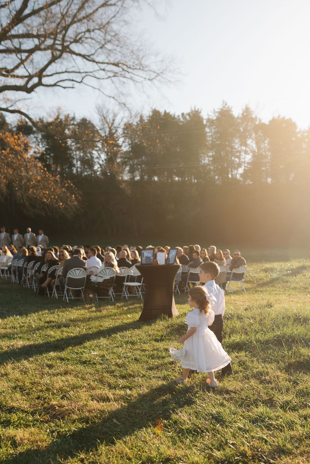 Outdoor wedding processional on grassy hill with guests seated in white chairs.