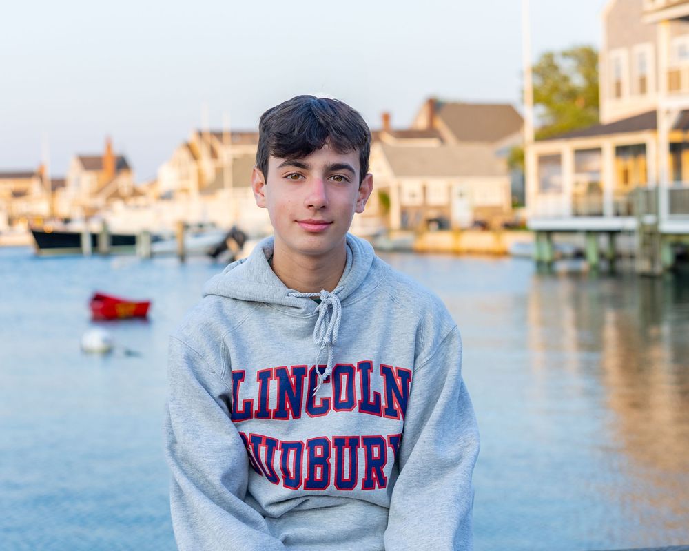 A teenager sits on a dock with boats and cottages visible in the background at the boat basin on Nantucket, MA by Lisa West Photography.