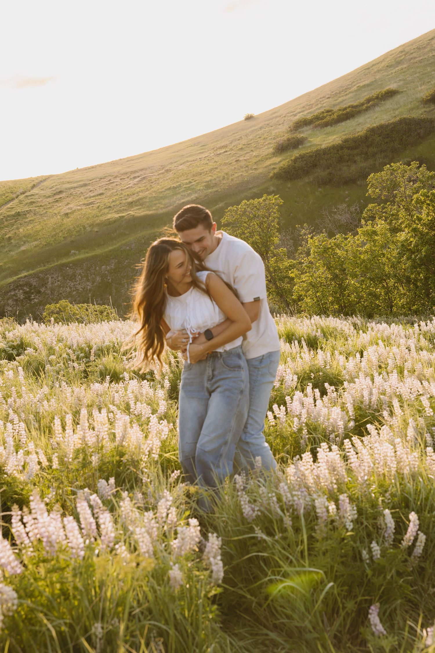 Bride in flowing white dress holds bouquet while standing on wooden deck surrounded by evergreen mountains at sunset.