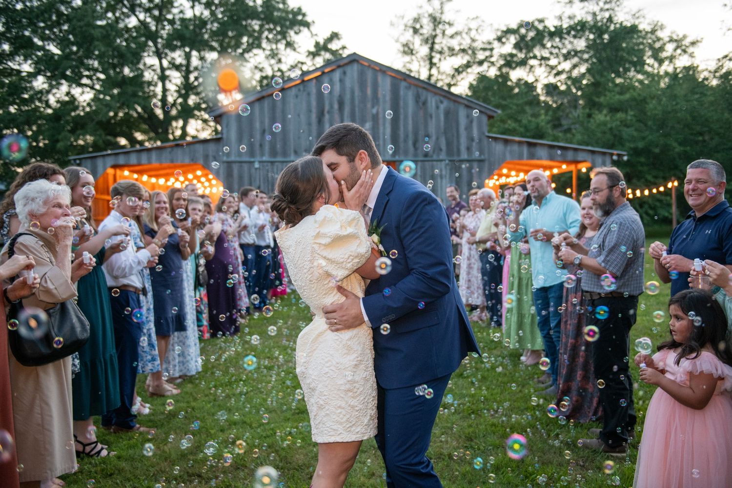 Outdoor wedding reception with string lights near a rustic barn as guests celebrate.