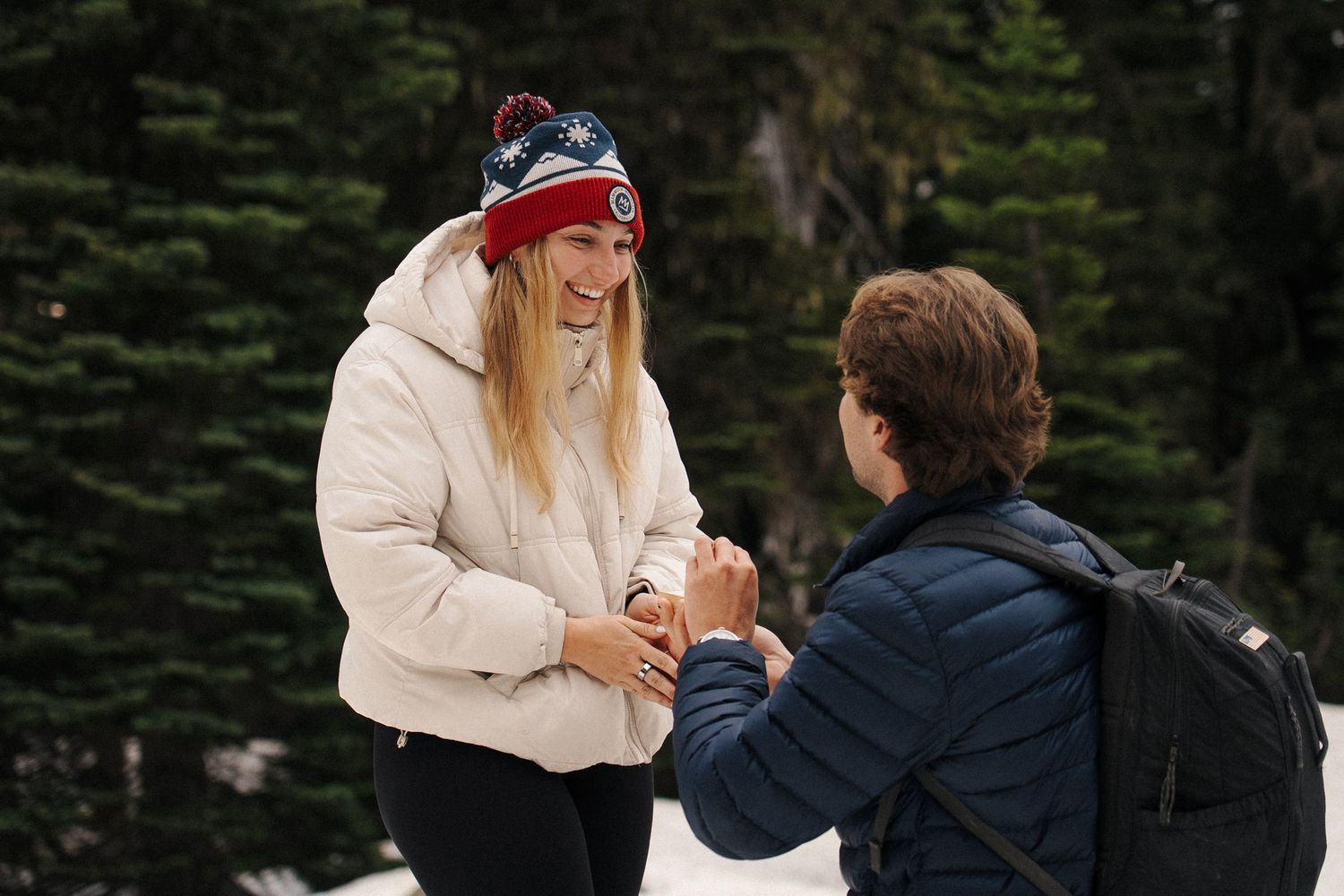 A winter outdoor proposal scene with two people in snowy weather wearing warm coats and winter accessories.