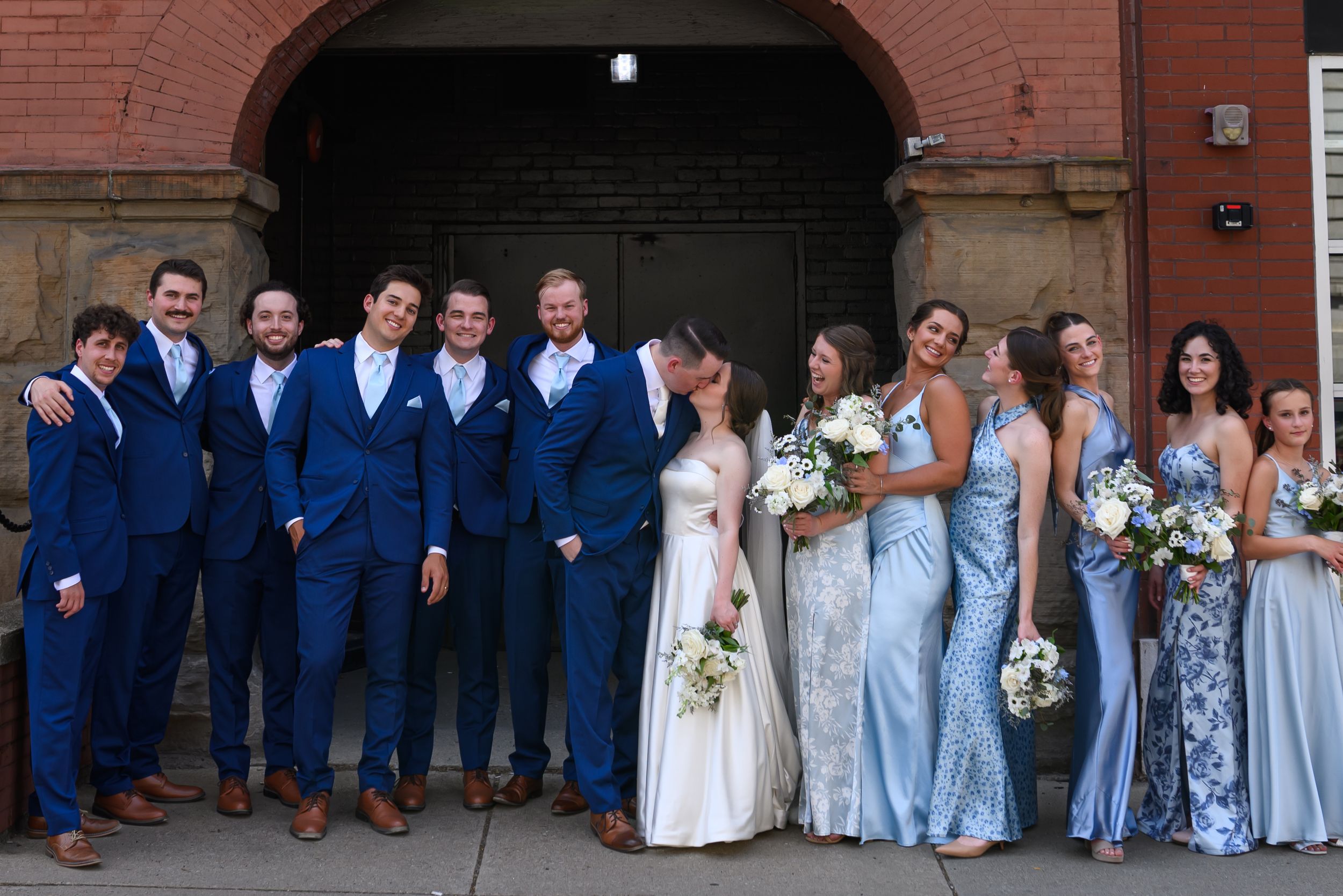 Wedding party members in navy suits and light blue dresses pose together for group photos in front of a brick building.