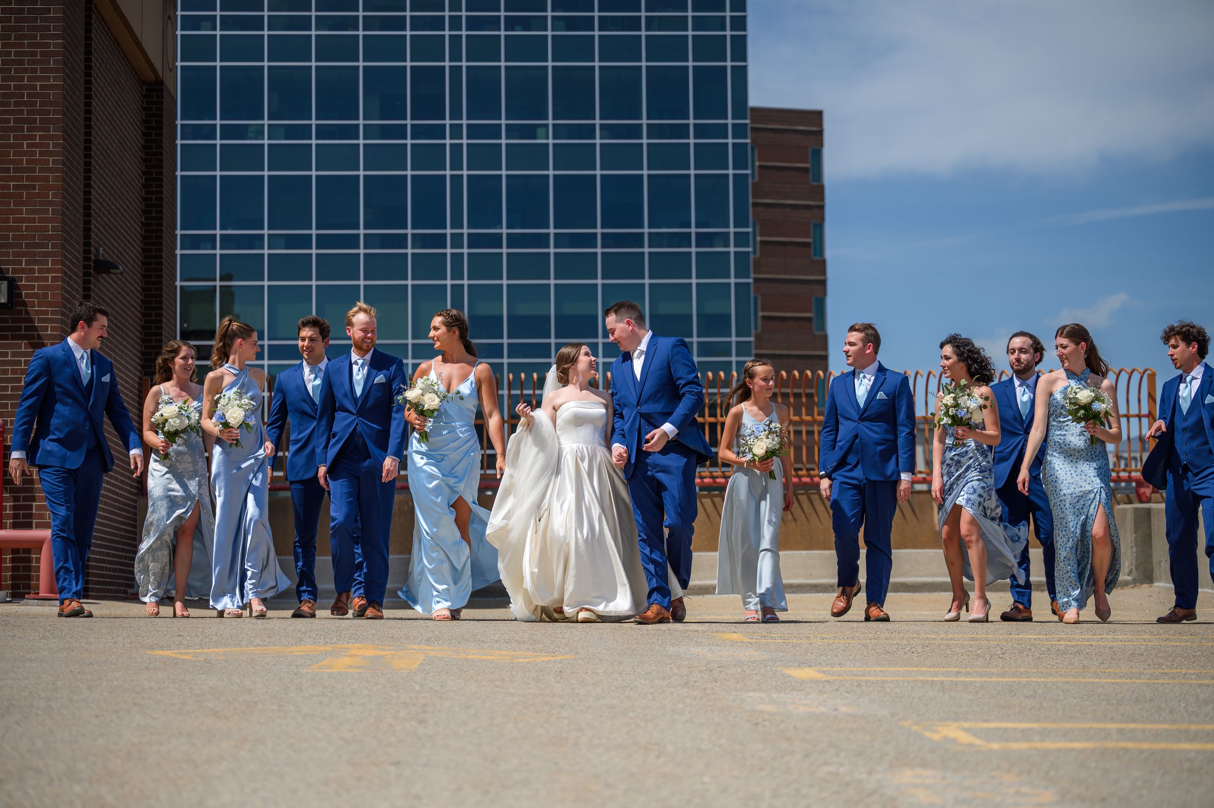 A wedding party in blue suits and elegant gowns poses together outside a modern glass building on a sunny day in Grand Rapids