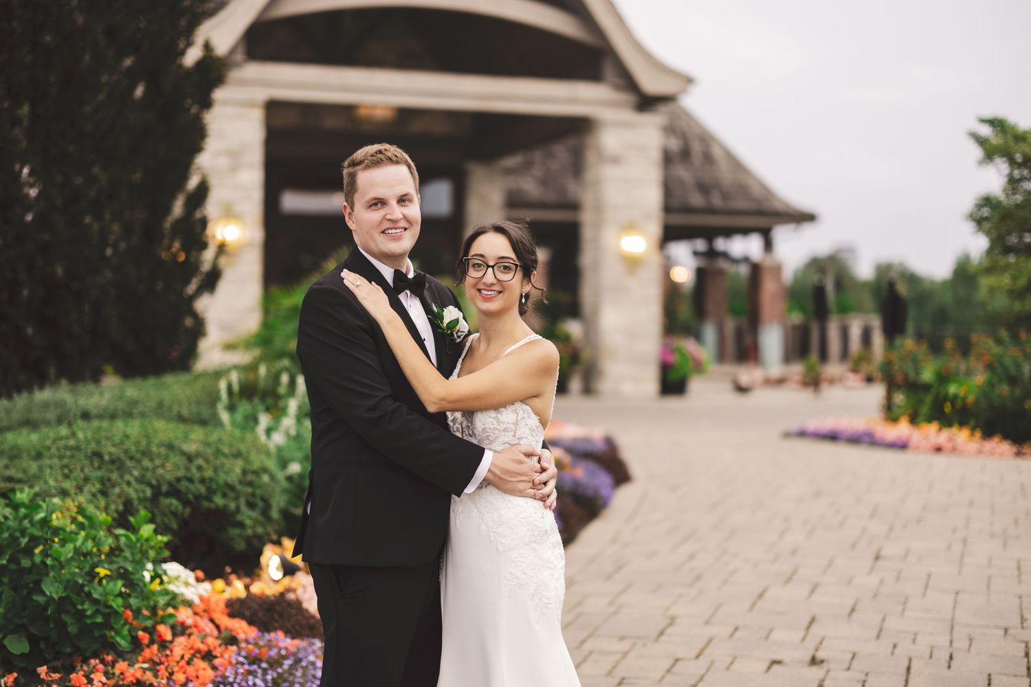 A bride and groom embrace on a stone walkway surrounded by colorful flowers and landscaping outside an elegant venue.