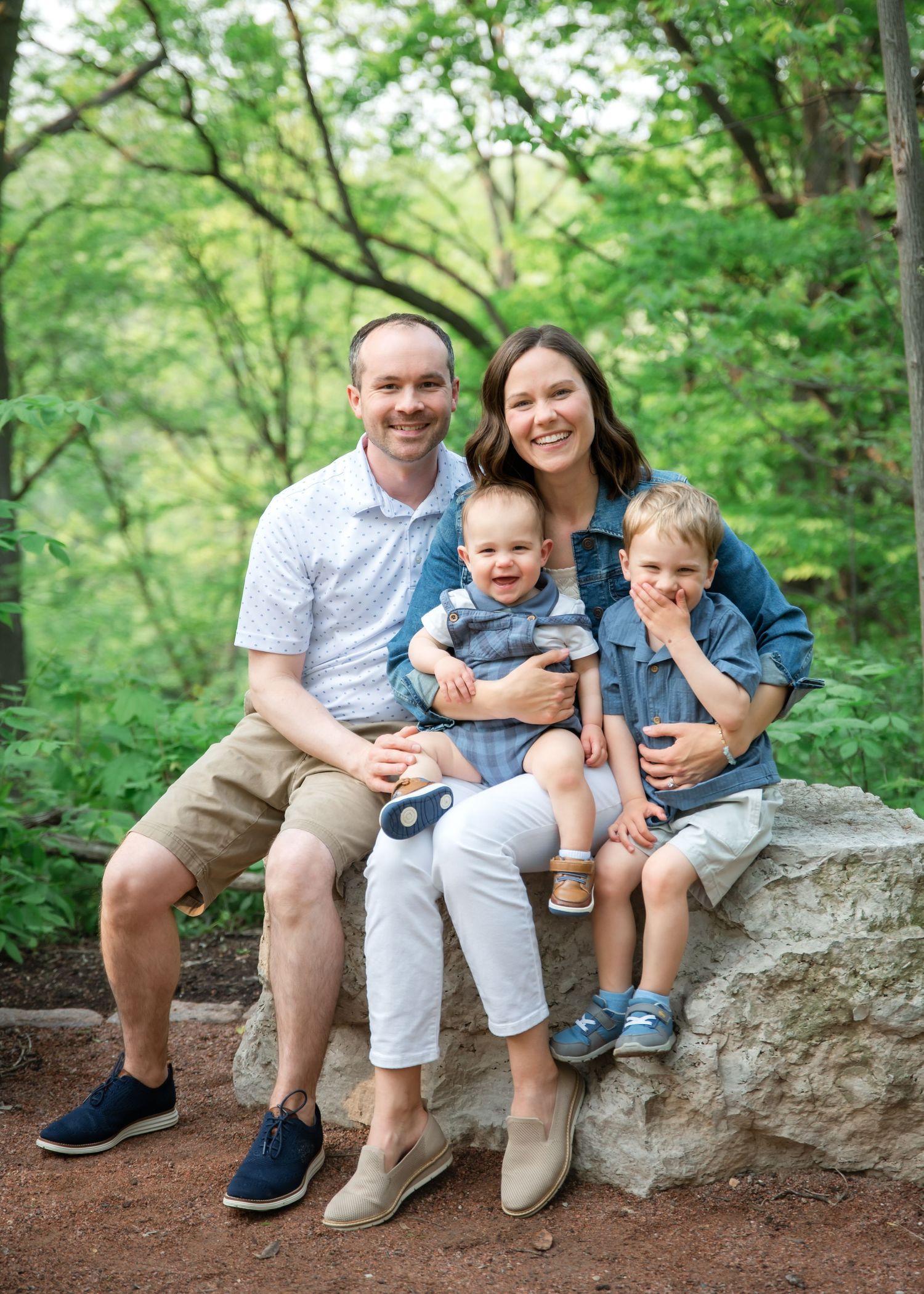 A family sits together on large rocks in a wooded outdoor setting during a photo shoot.