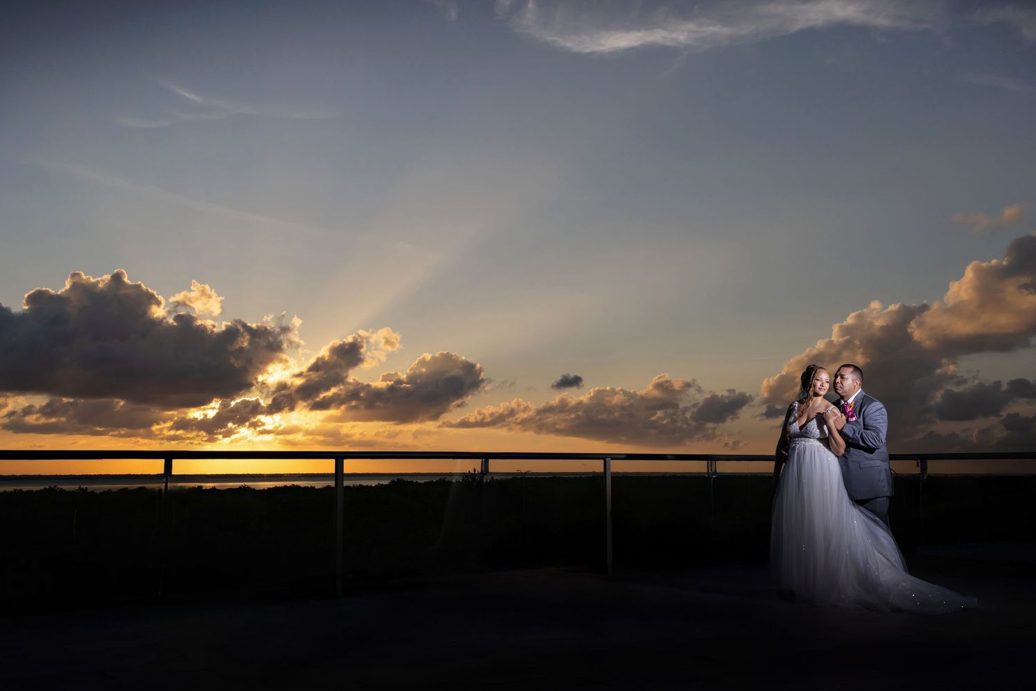 A couple in wedding attire stands together against a dramatic sunset sky with golden clouds over water at Planet Hollywood Cancun.