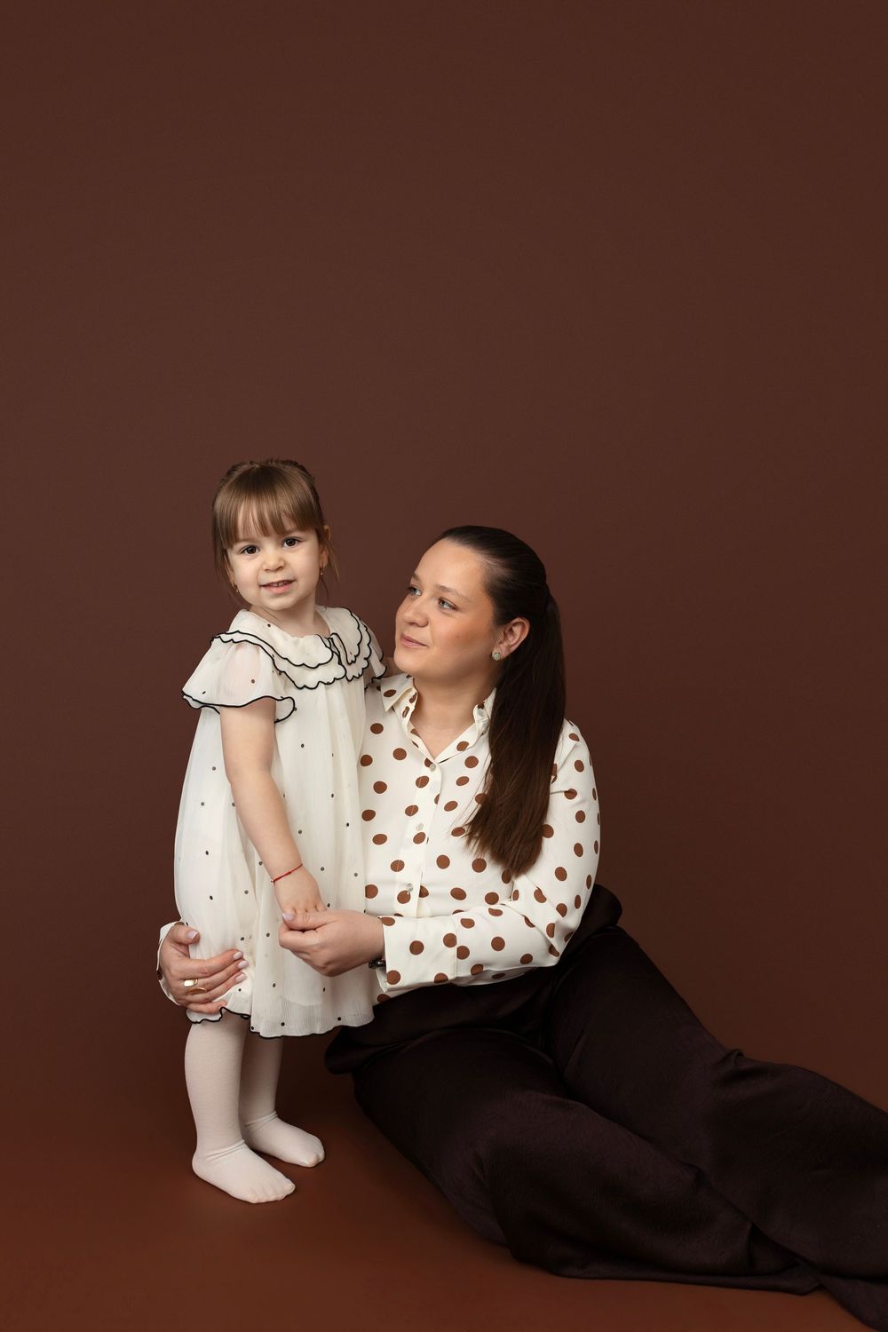 A parent and child wearing white and polka dots pose together against a brown backdrop in a studio setting.