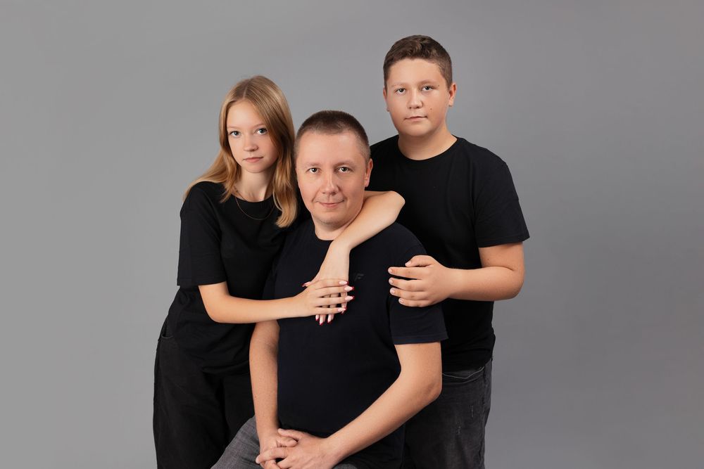 A family wearing matching black outfits poses together against a grey studio backdrop for a portrait.