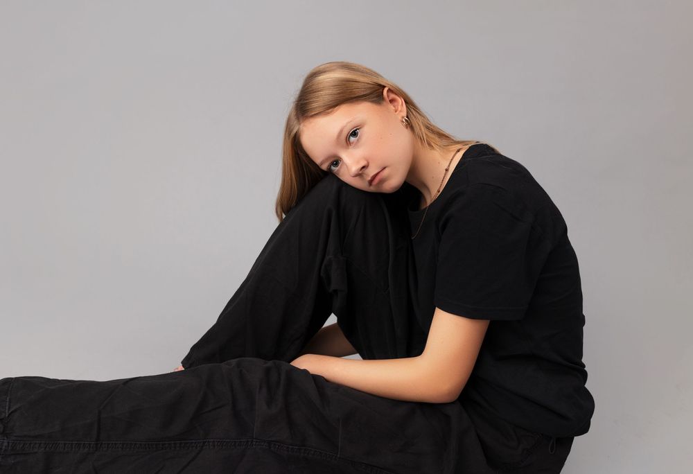 Person in all black attire sits against gray background while leaning head to the side in contemplative pose.