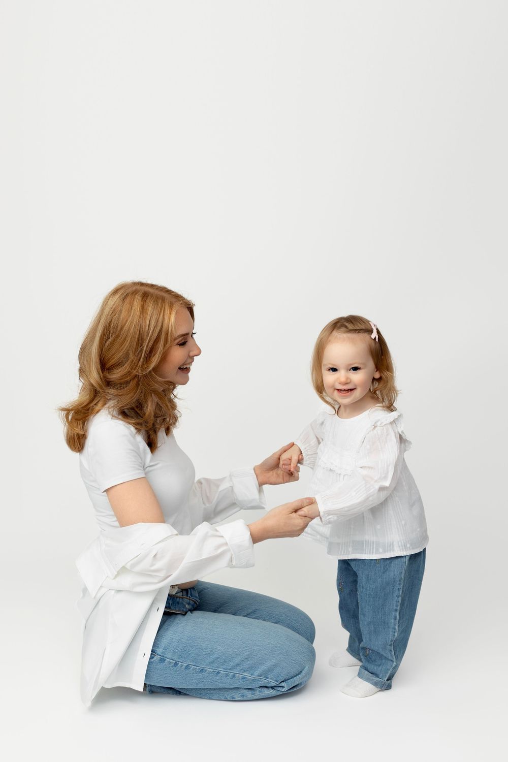 Mother and toddler wearing white tops and jeans share a playful moment on a light background.