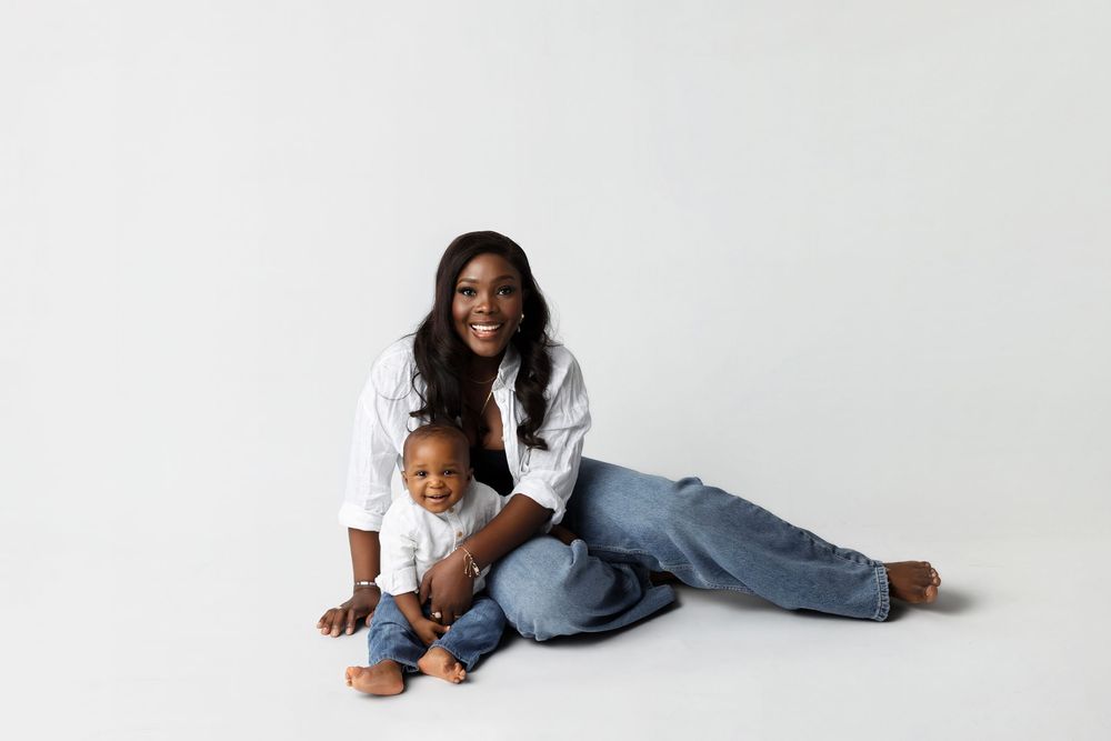 A smiling Black mother in white shirt and jeans sits on white floor with her happy baby boy.