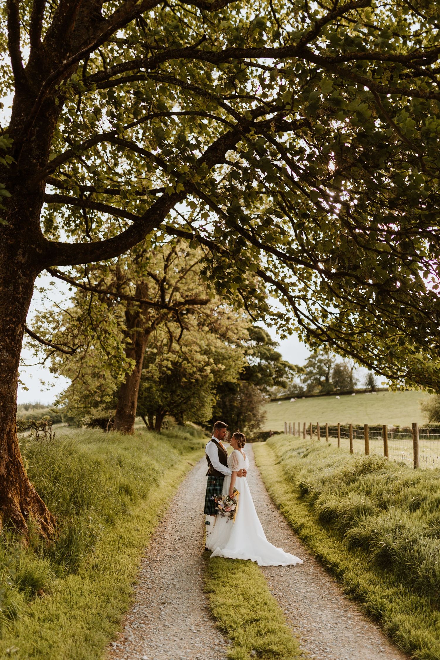 Couple walking along a scenic tree-lined dirt path on a sunny summer day in countryside setting.