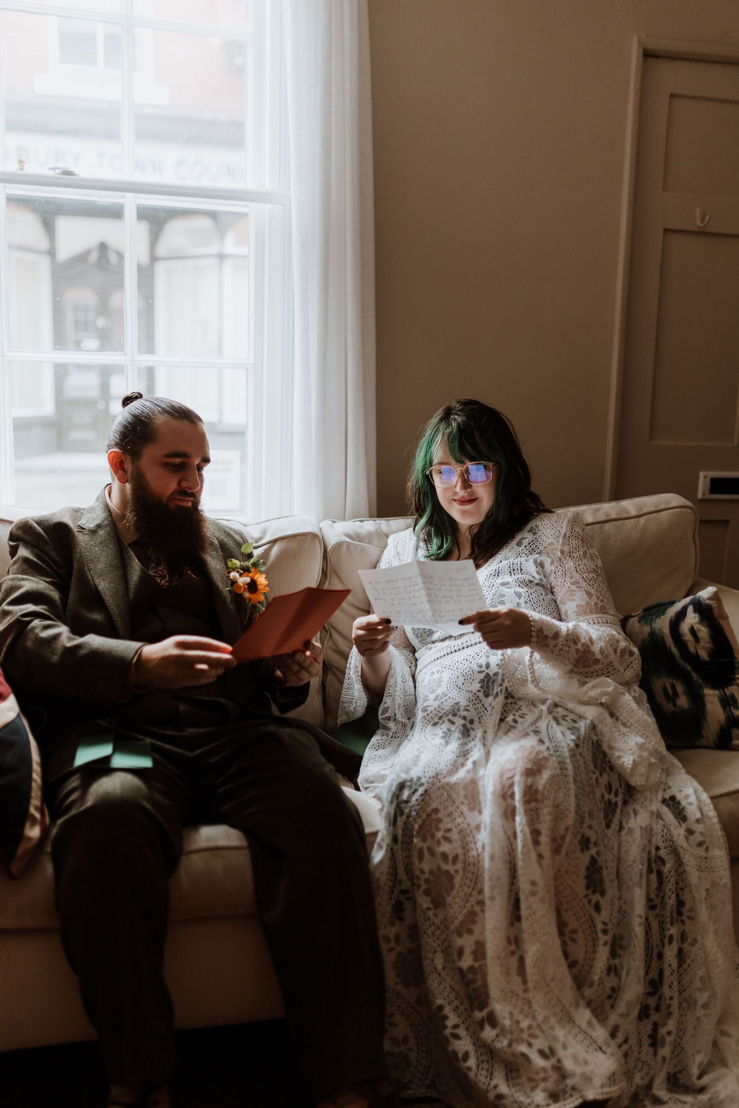 A couple reads together on a couch by a window, with the person on the right wearing an elegant lace dress.