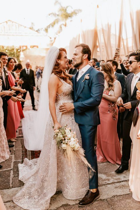 Couple shares intimate moment during sunset wedding reception with guests in coral dresses.