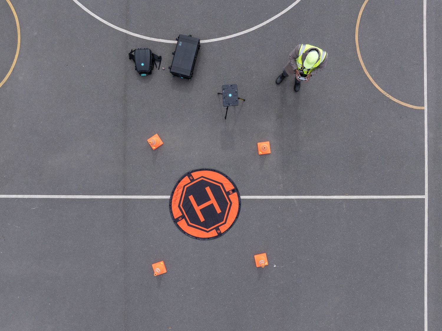 Aerial view of drone landing platform with orange helipad marking and scattered cones on concrete surface.