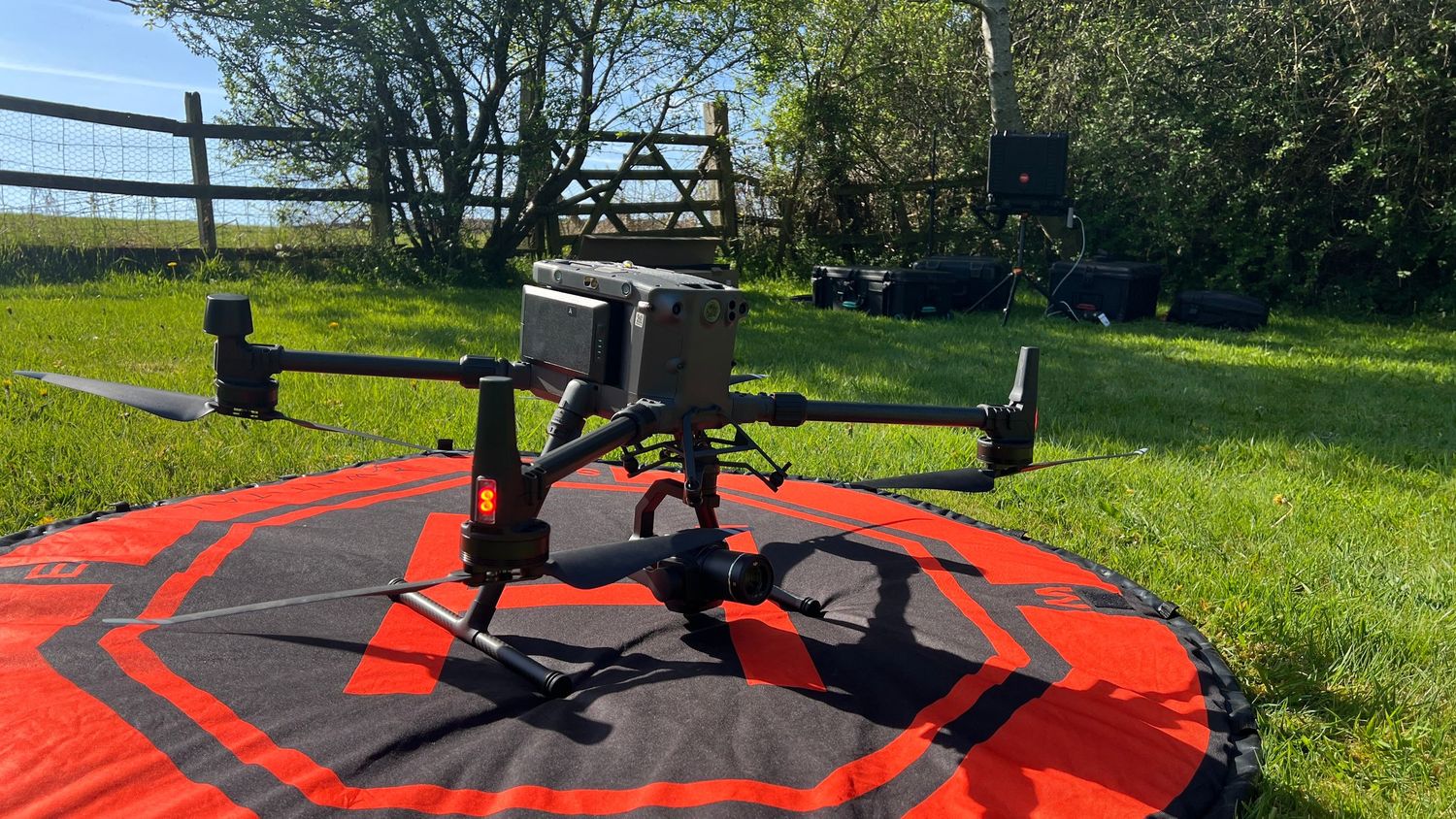 A black drone rises against a sunny sky during flight operations in a grassy field.
