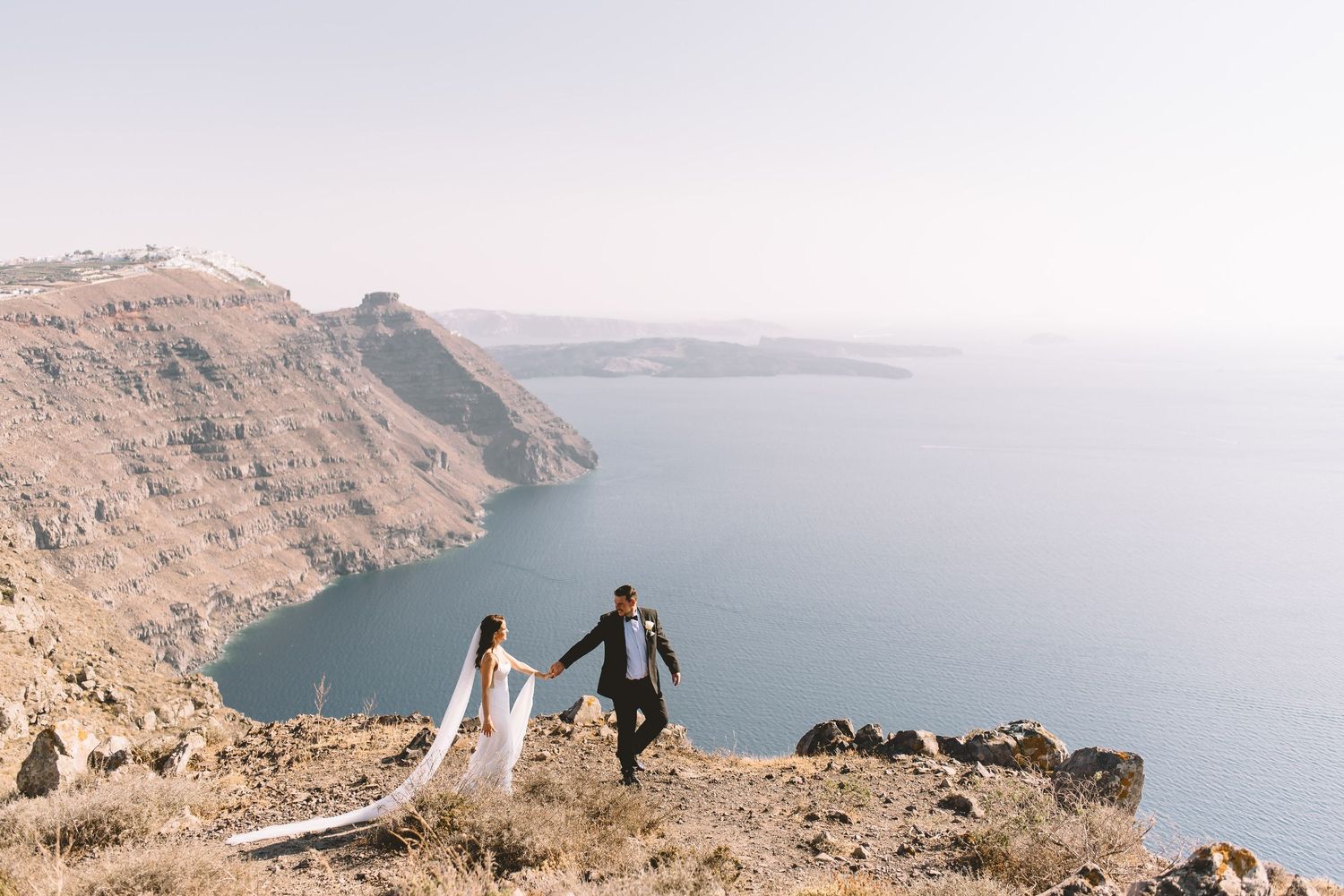 Couple in wedding attire standing on cliff edge overlooking blue Mediterranean waters in scenic Santorini landscape.