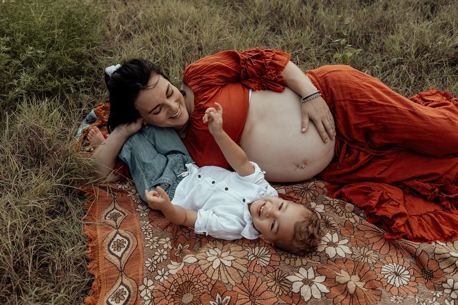 A maternity photoshoot in a grassy field shows a woman in a flowing red dress lying gracefully among wildflowers.