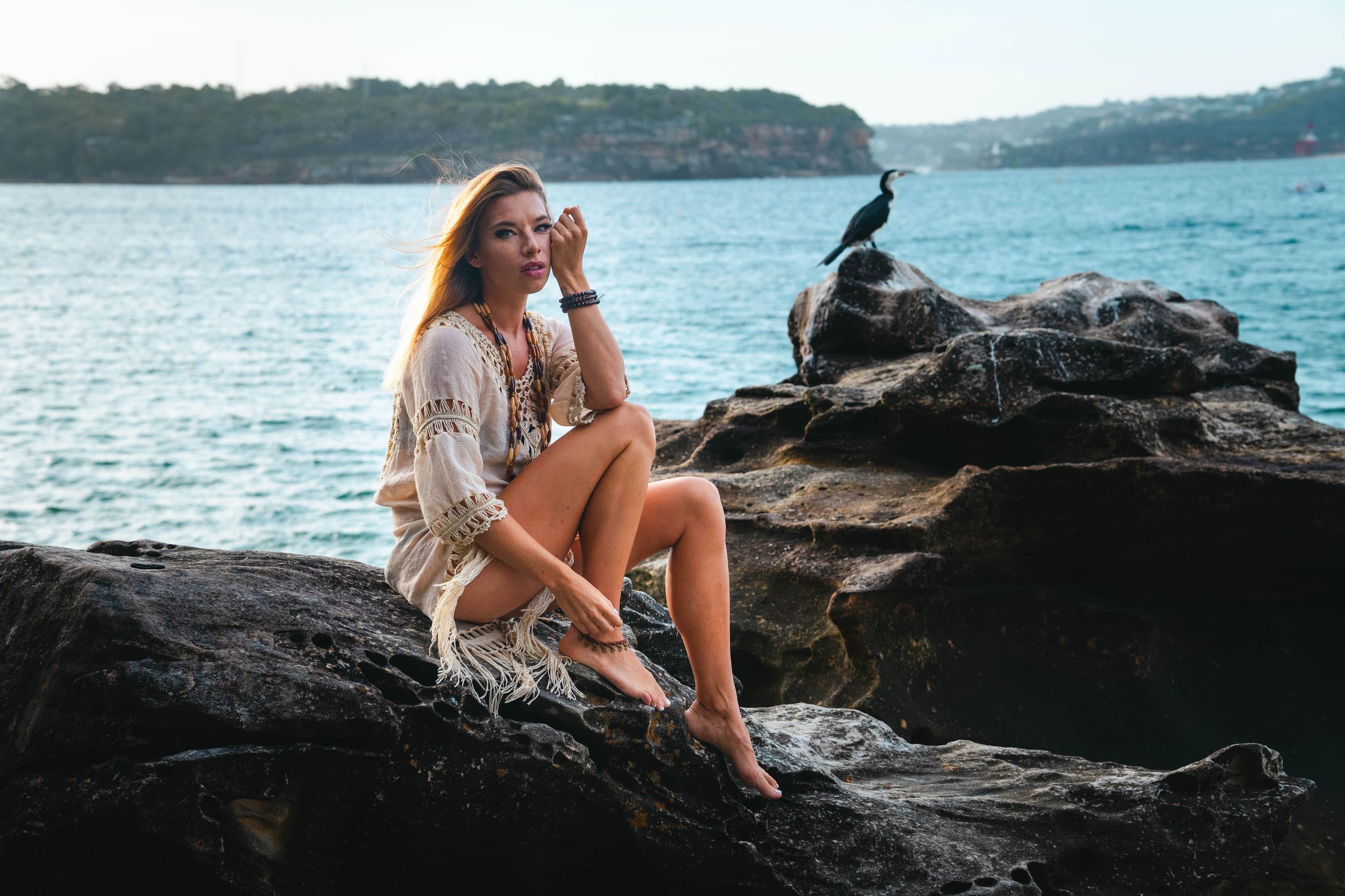 A figure in a crochet coverup sits on coastal rocks by blue waters while a cormorant perches nearby.
