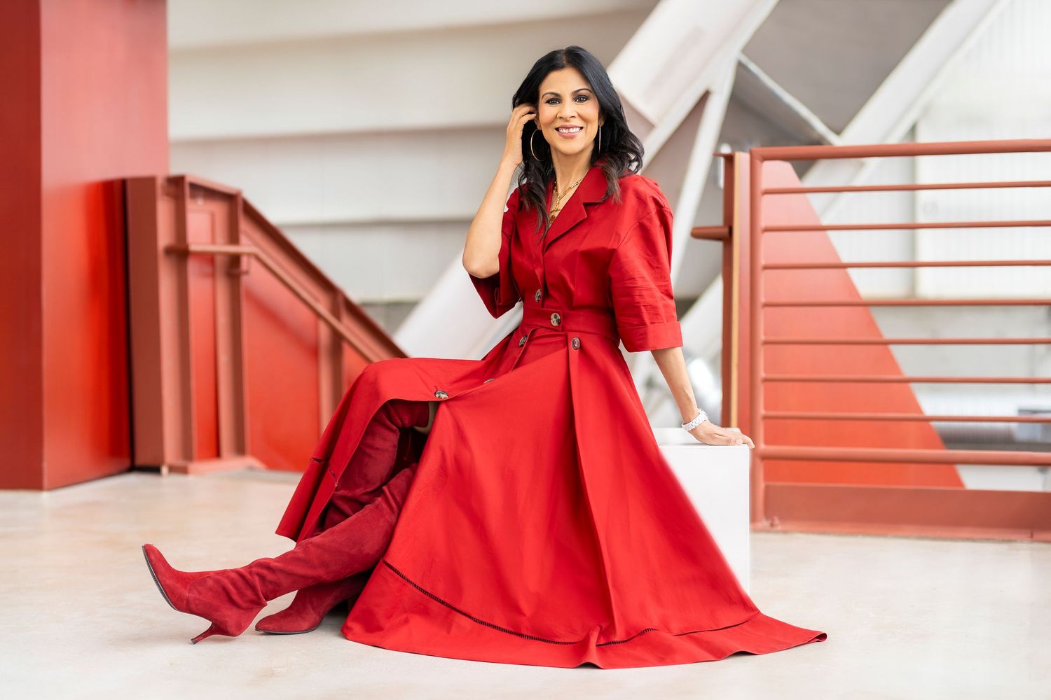 Woman in flowing red dress and boots seated on modern architectural steps.