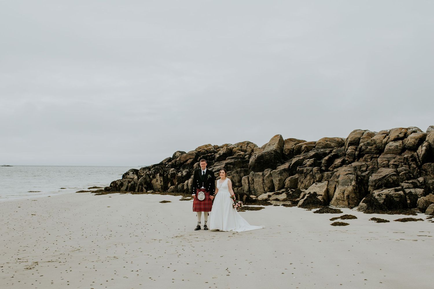 Person in kilt standing near rocky shoreline on cloudy beach day.