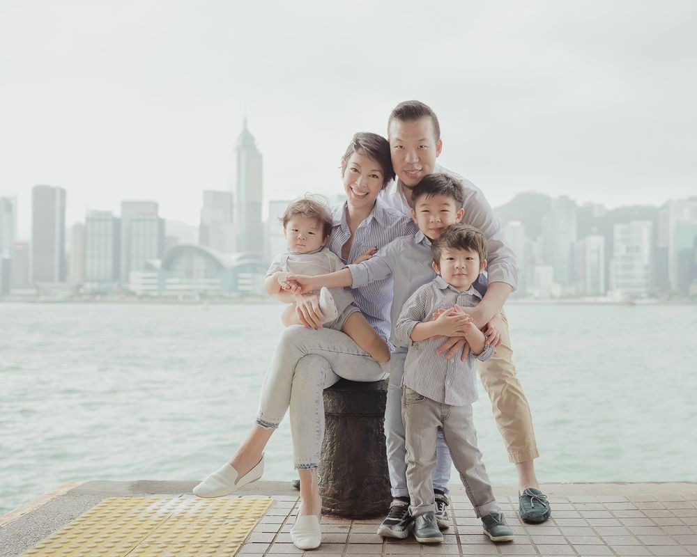 A family in casual attire sits together and poses for photos along a waterfront with a city skyline in the background.