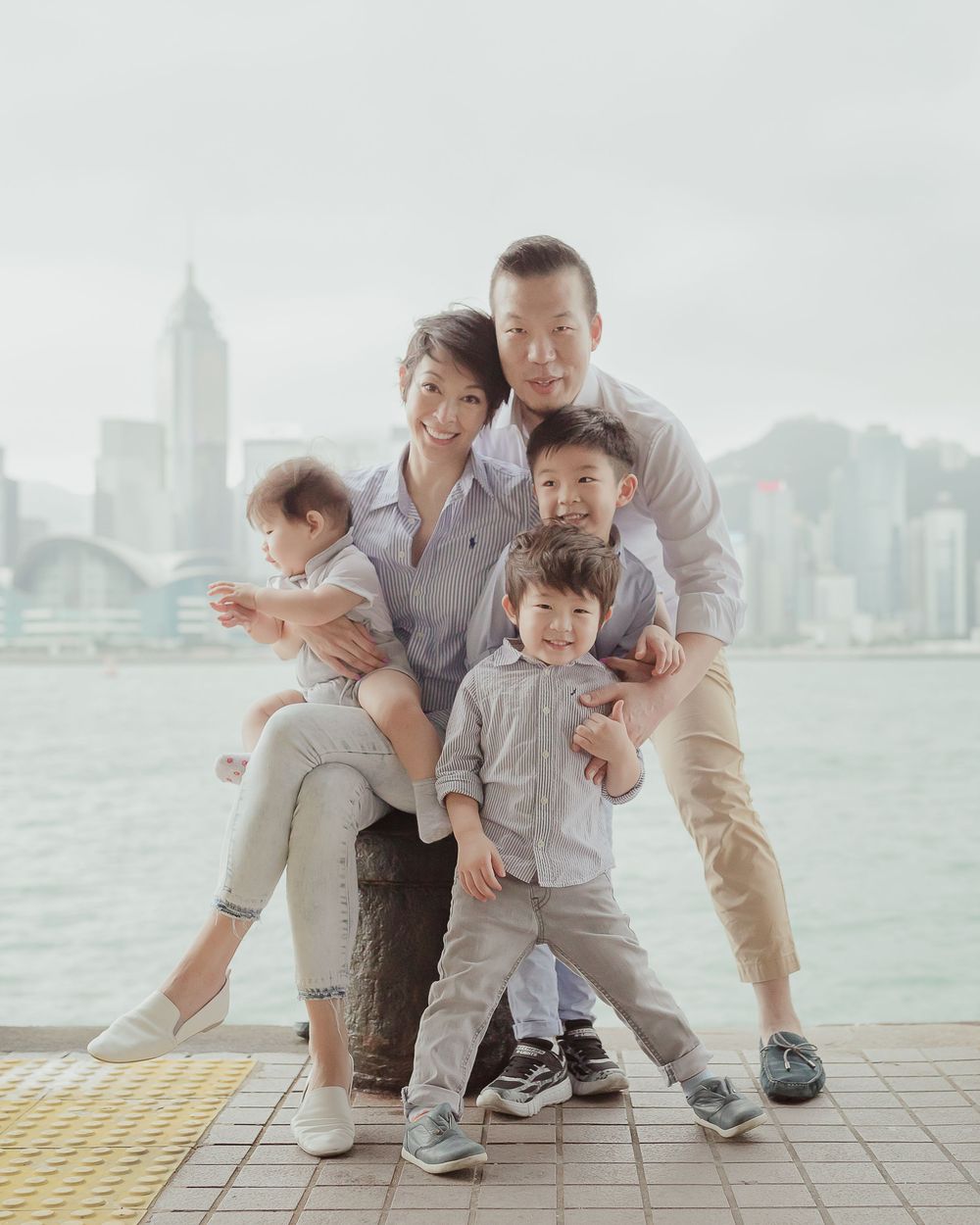 A family shares a tender moment together on a waterfront promenade overlooking Hong Kong's Victoria Harbor in matching outfits.