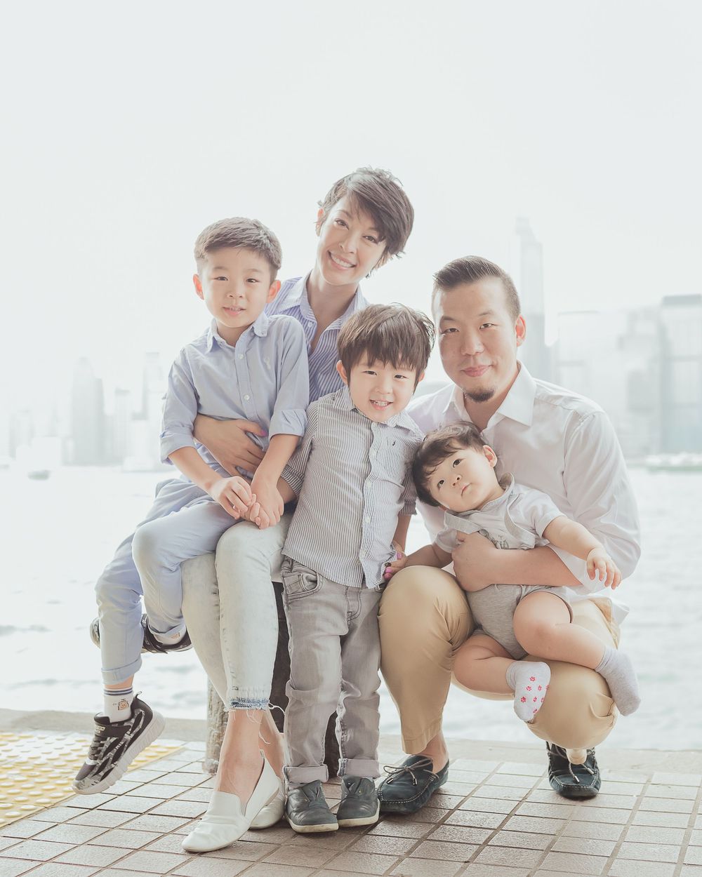Family enjoying playful moment while posing together on outdoor paved walkway with cityscape in background.