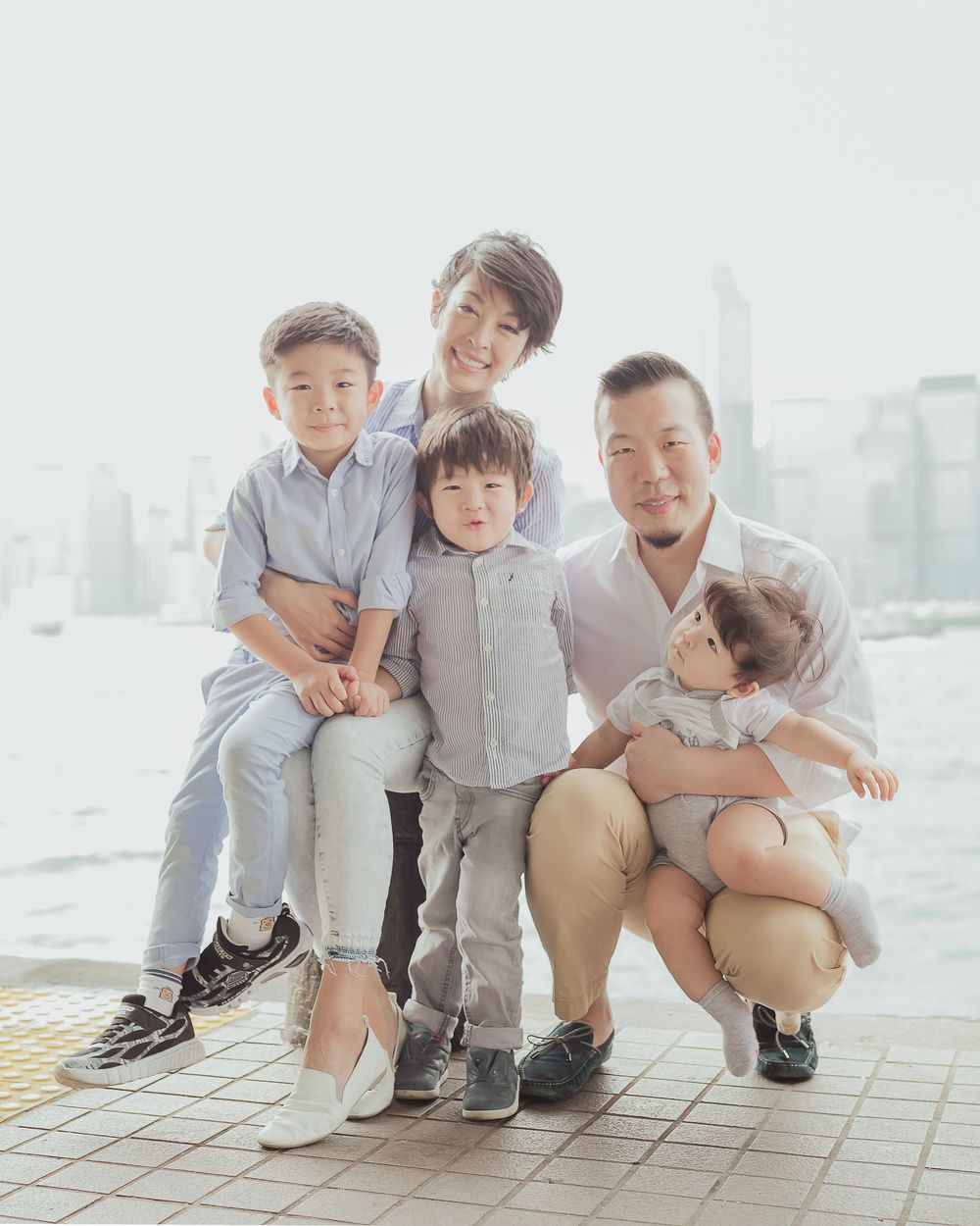 Family dressed in light colors poses together against city skyline backdrop on tiled outdoor viewing area.
