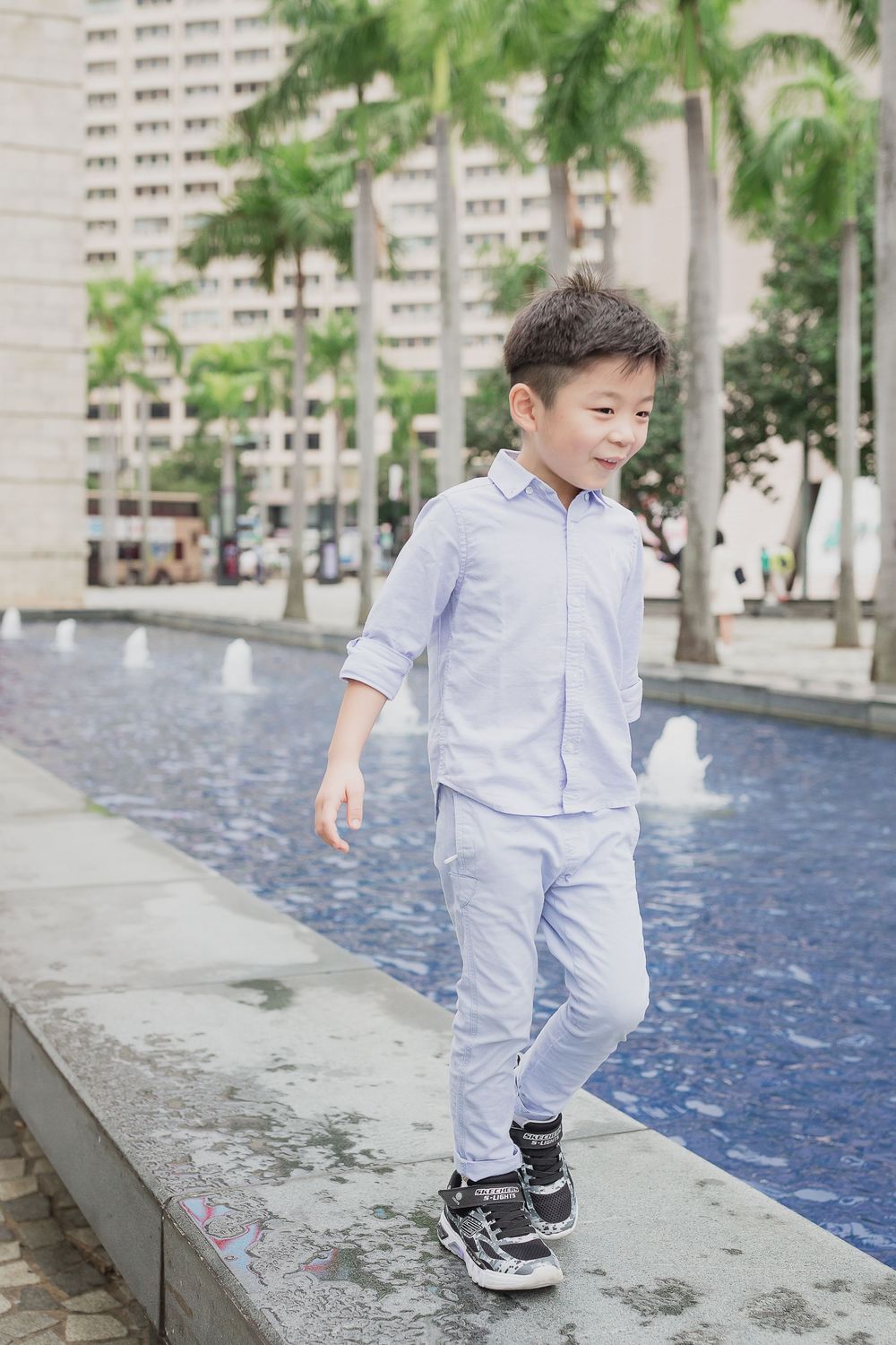 A young child in a light gray outfit plays along the edge of a decorative water feature surrounded by palm trees.