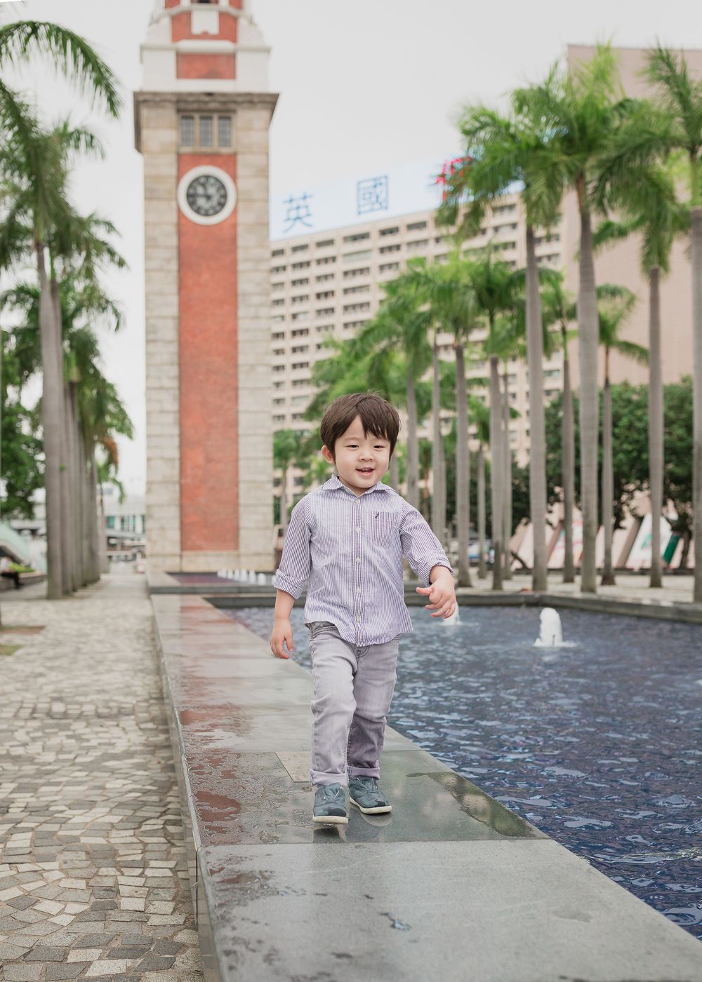 A child plays and runs along the edge of a blue reflecting pool with palm trees and a clock tower in the background.