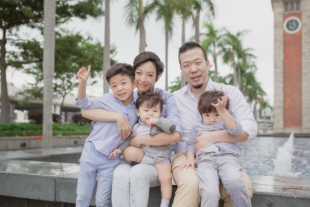 A family wearing matching grey outfits poses together cheerfully on outdoor steps near palm trees.