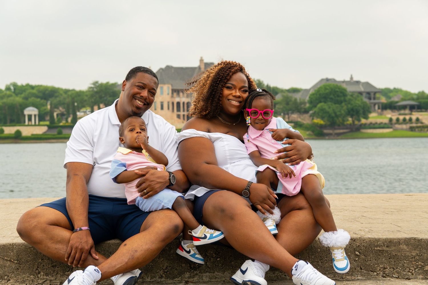 A family in matching white outfits sits together at the beach, smiling and holding two babies near the water.
