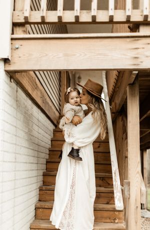 Mother in white dress holding baby on vintage wooden staircase.