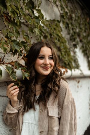 Young woman in beige cardigan poses against ivy-covered wall.