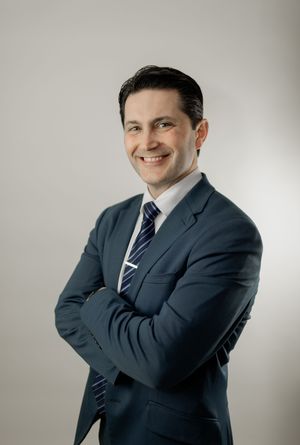 Professional business portrait of someone in a navy suit and striped tie posing confidently against a light background.